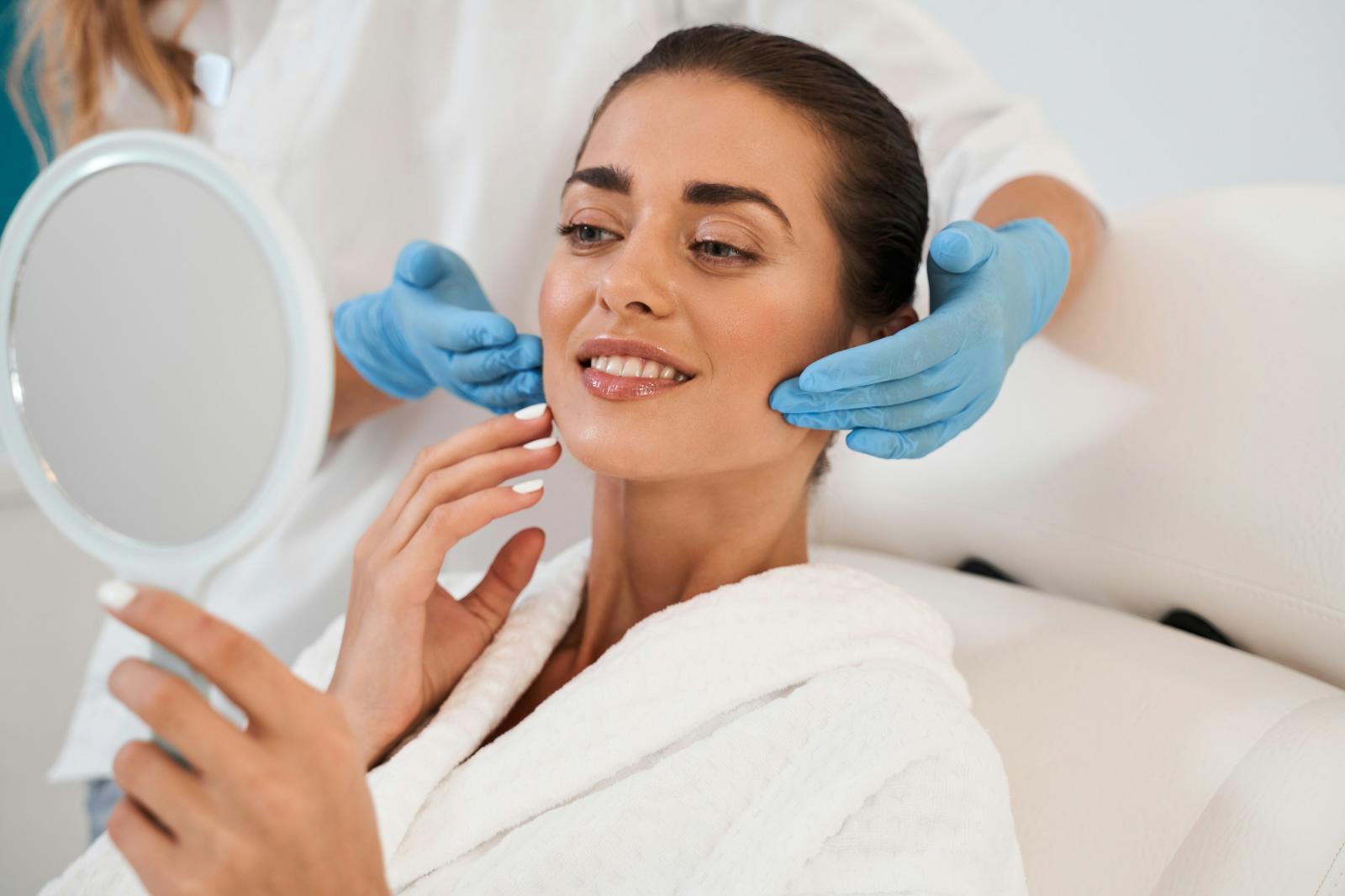 Client in a white robe looking into a hand mirror while an aesthetics provider in blue gloves performs a facial skin assessment.